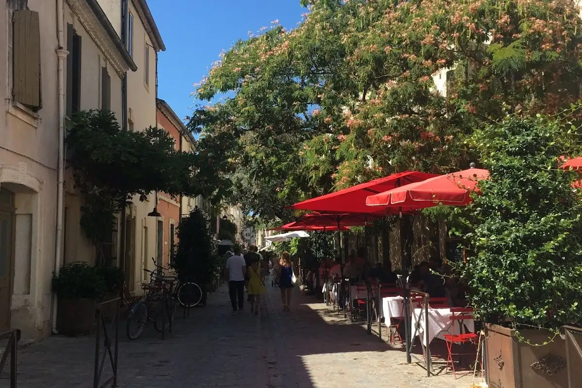 Rue étroite et pittoresque avec des cafés sous des parasols rouges, des arbres verdoyants et des passants par une journée ensoleillée.