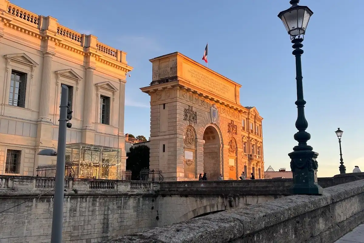 Arche en pierre historique et bâtiments illuminés par la lumière dorée du coucher du soleil, avec un lampadaire et un pont au premier plan.
