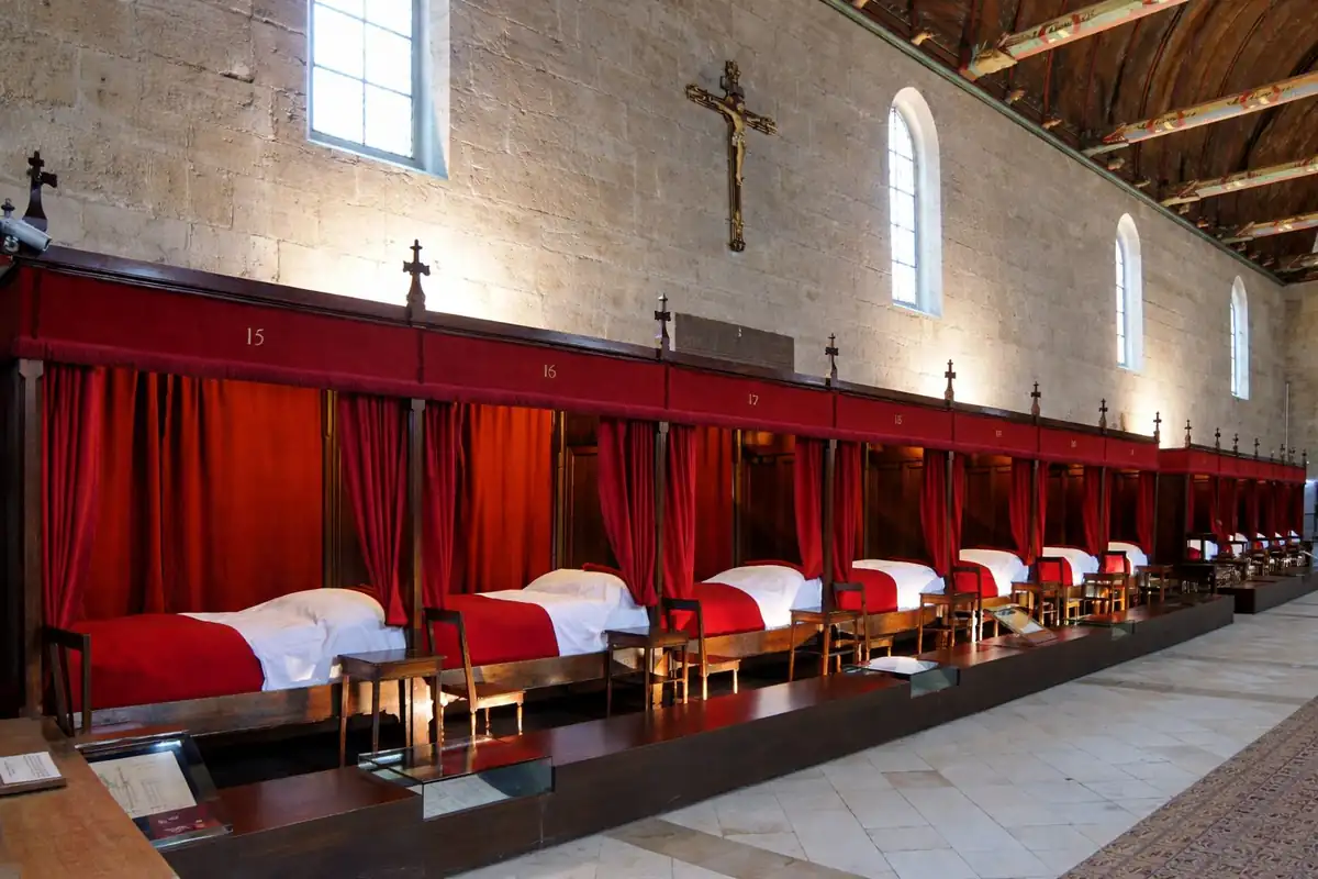 Grande salle des malades des Hospices de Beaune : lits alignés à rideaux rouges sous une charpente sombre médiévale