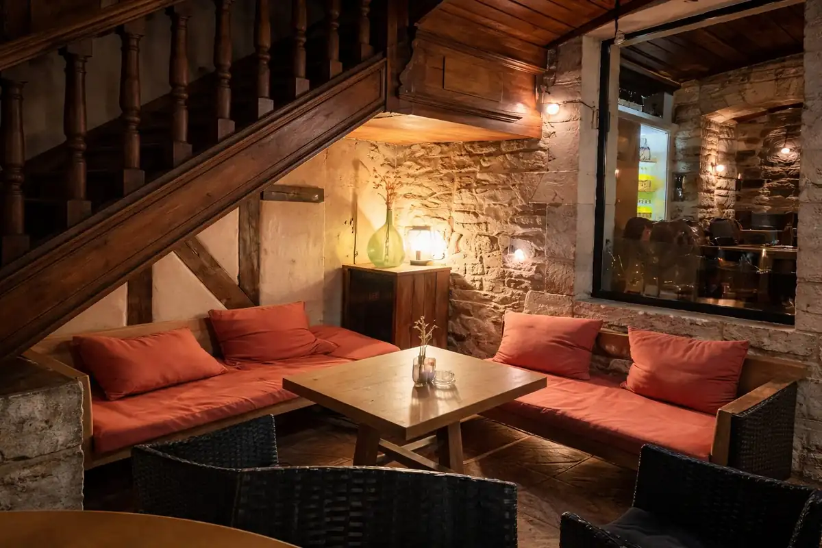 Intérieur chaleureux du Bout du Monde : salon en pierre avec canapés aux coussins rouges et escalier en bois sous une lumière dorée